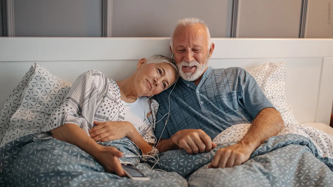 Couple âgé allongé confortablement au lit, écoutant de la musique ensemble avec des écouteurs.