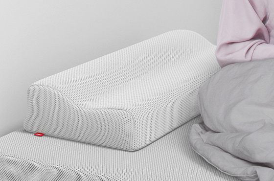  Oreiller ergonomique BODYGUARD® posé sur un lit, à côté d’une personne assise sous une couette grise.