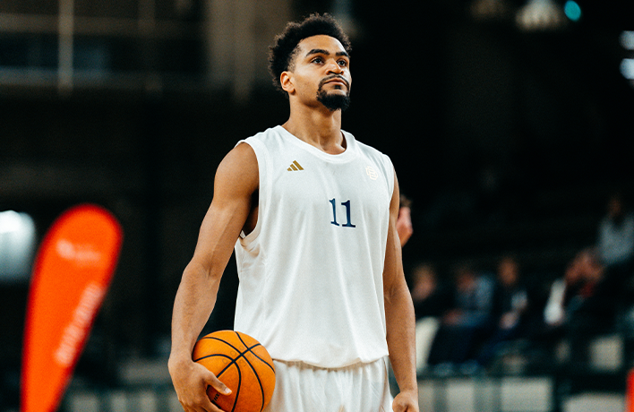 Joueur de basketball en tenue rouge BRVS n°3 tenant un ballon, debout devant un panneau d’affichage lumineux en salle.