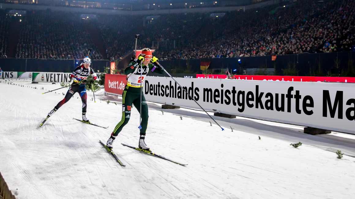 Foto: Vereinzelte Personen auf Skiern hintereinander auf einer Laufbahn im Stadion. Auf der Stadionbande steht in großen Lettern: Deutschlands meistgekaufte Matratze.