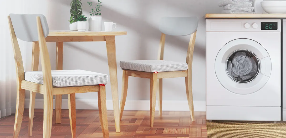 Deux coussins de chaise BODYGUARD® posés sur des chaises en bois clair autour d’une table dans une cuisine. À droite, un lave-linge affiche 60 °C, suggérant que les housses des coussins sont lavables.