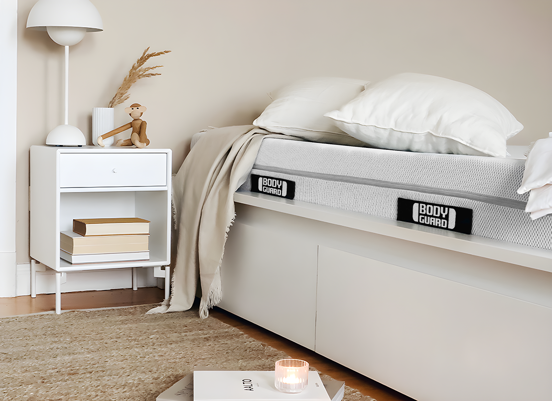 Elément d'une chambre à coucher avec un lit visible sur lequel repose un matelas BODYGUARD. A côté du lit se trouve une table avec une lampe de chevet.