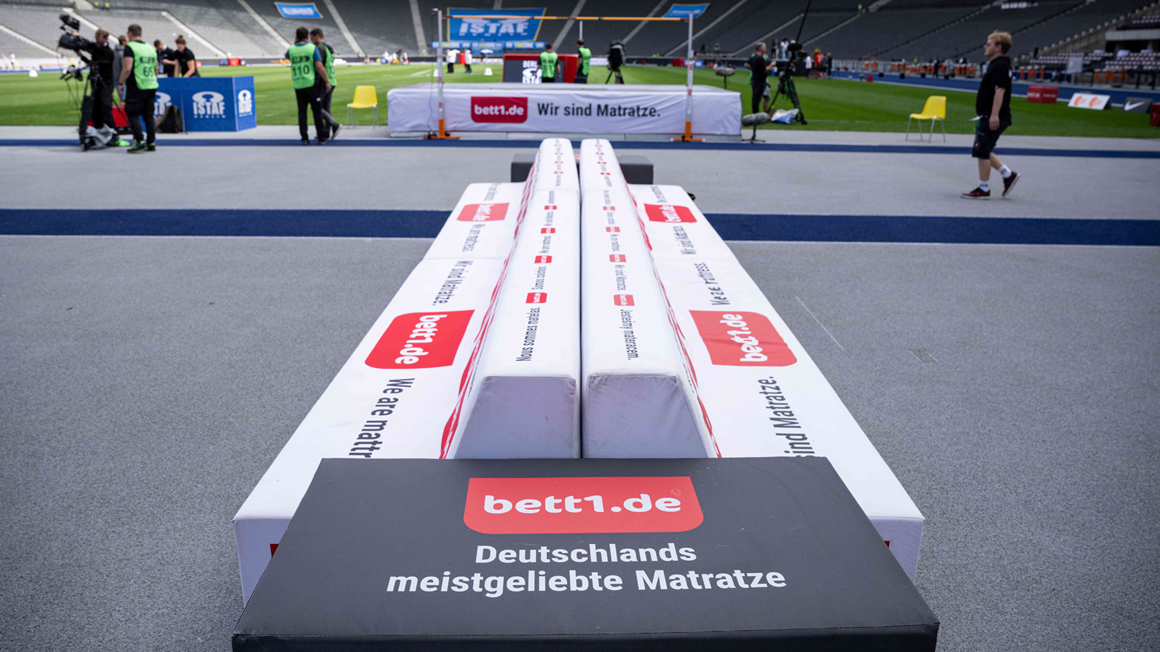 Foto vom Spielfeldrand in einem Stadion, über eine Spielendenbank mit bett1 Logo auf Leichtathletik-Stationen in der Rasenmitte.