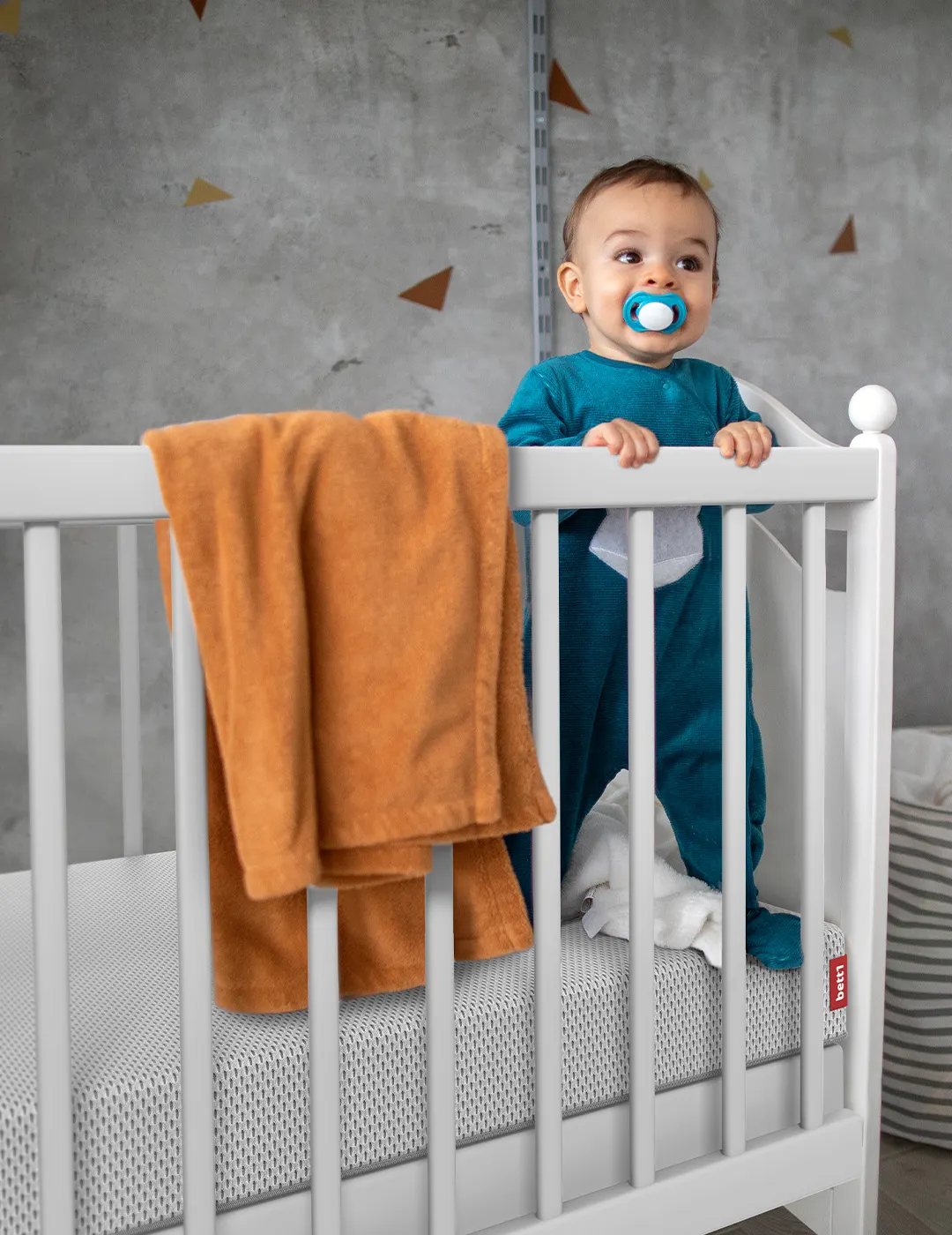 Baby steht in Gitterbett auf der BODYGUARD® Kindermatratze. Text: „Die verstärkte Außenseite finde ich besonders gut, da das Kind sicher am Rand stehen kann.“ Lisa-Marie aus Niedernwöhren