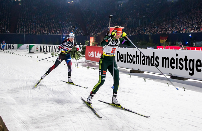 Compétition de biathlon avec deux athlètes en pleine course sur neige, devant une foule en tribunes. Les panneaux publicitaires affichent notamment le logo bett1.de et la mention « Deutschlands meistgekaufte Matratze.