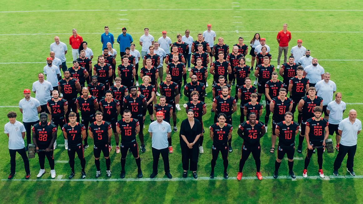 Foto: Das Team der Berlin Braves in mehreren Reihen auf dem Rasen. Die Spielenden in schwarz-roten Trikots, Management und Stuff drumherum in weißen Oberteilen.