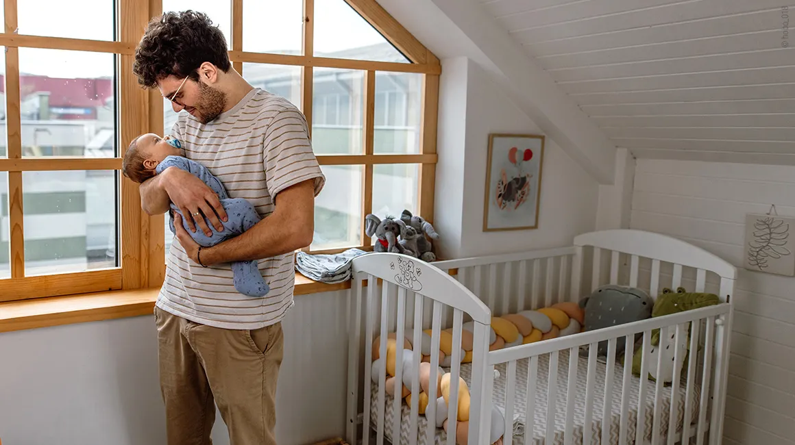 Jeune papa berçant son bébé dans une chambre lumineuse avec un lit à barreaux.