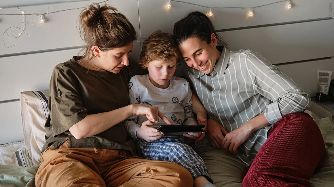 Famille composée de deux mamans et leur enfant, souriants en regardant une tablette depuis leur lit.
