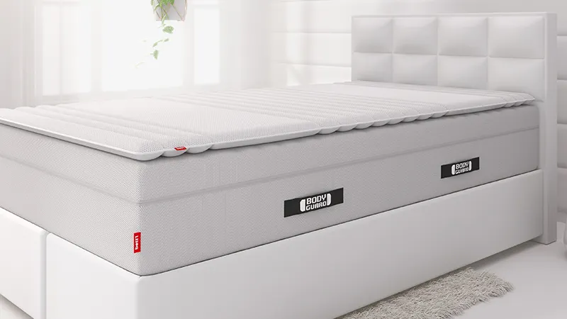 Auf einem weißen Boxspringbett liegt die extra hohe BODYGUARD Matratze und darauf der Topper 140x200 cm.