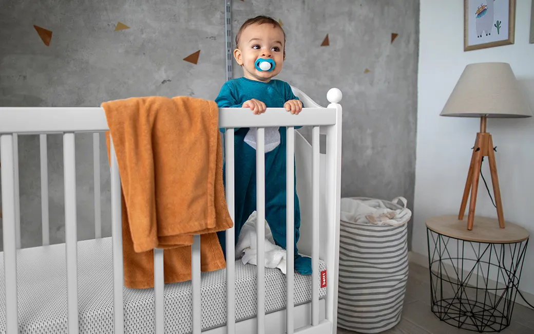 Baby steht in Gitterbett auf der BODYGUARD® Kindermatratze. Text: „Die verstärkte Außenseite finde ich besonders gut, da das Kind sicher am Rand stehen kann.“ Lisa-Marie aus Niedernwöhren