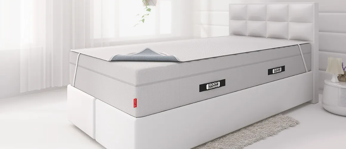 Lit boxspring moderne avec matelas BODYGUARD® Boxspring et surmatelas gris partiellement replié, dans une chambre lumineuse au style épuré.