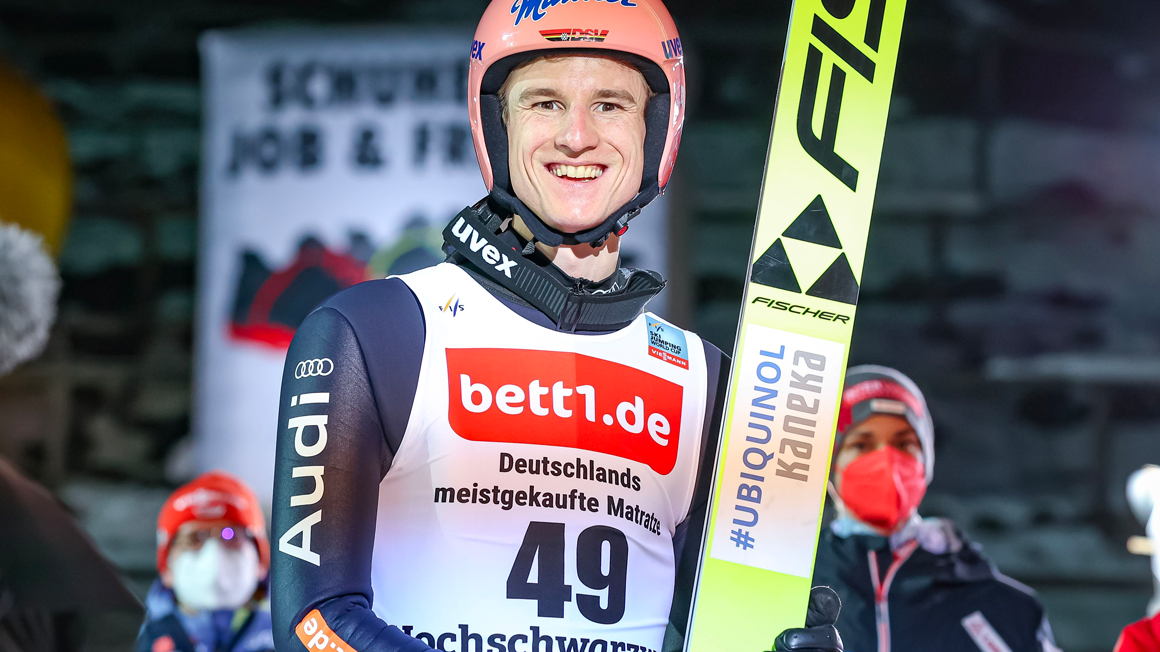 Skieur souriant en tenue de saut à ski avec casque rose, tenant ses skis. Son dossard porte le logo bett1.de.