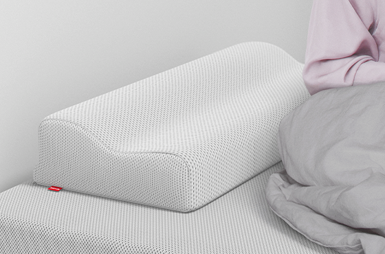  Oreiller ergonomique BODYGUARD® posé sur un lit, à côté d’une personne assise sous une couette grise.