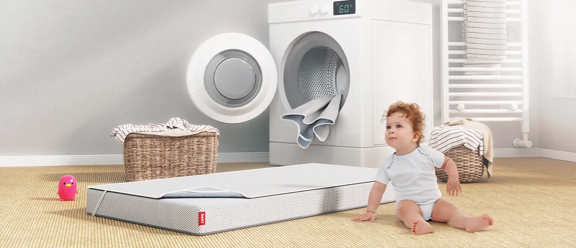In einem Badezimmer sitzt ein Kleinkind vor einer Waschmaschine. Daneben liegt die SuperBreeze Kindermatratze auf dem Boden. Sie ist mit dem BODYGUARD Nässeschutz bezogen.