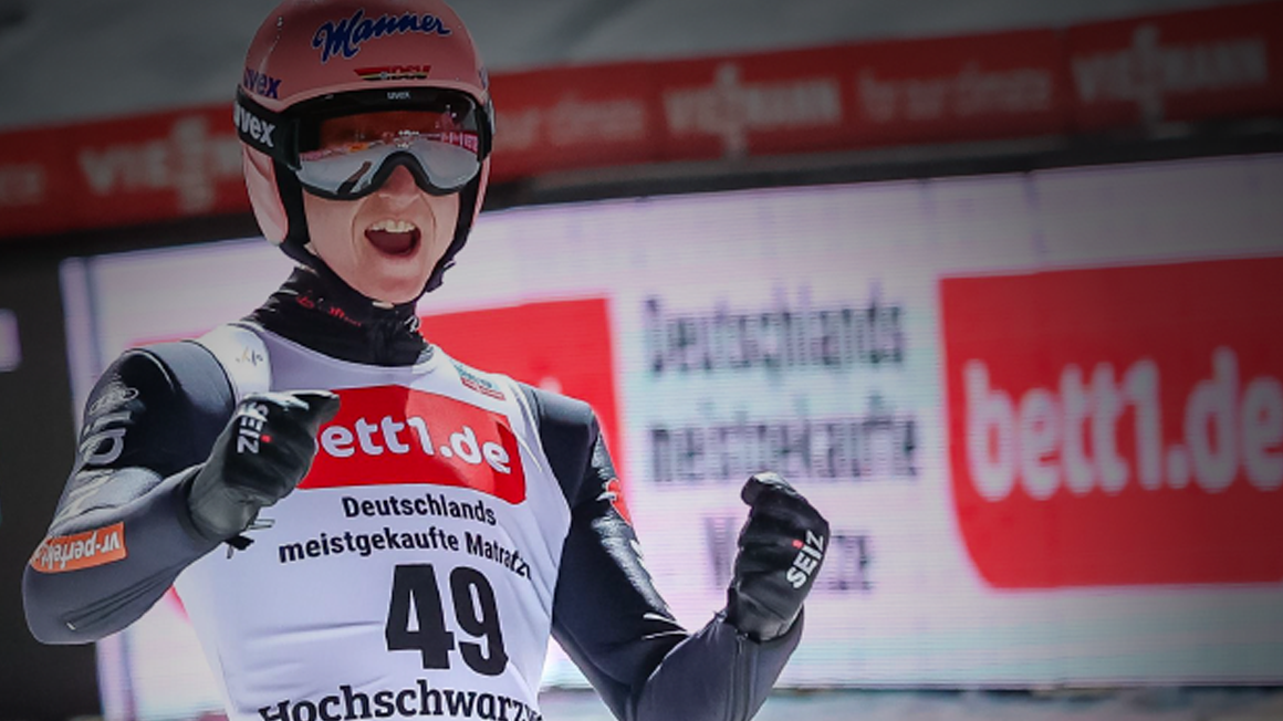 Athlète de saut à ski en tenue de compétition, levant les poings en signe de victoire. Logo bett1.de bien visible sur le dossard et en arrière-plan.
