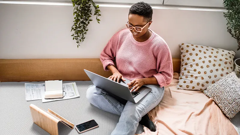 Jeune femme travaillant ou étudiant sur un lit, entourée de papiers et de coussins.
