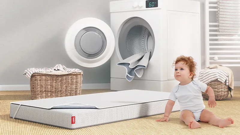 Bébé assis près d’un matelas pour enfant BODYGUARD®, devant une machine à laver ouverte dans une buanderie.