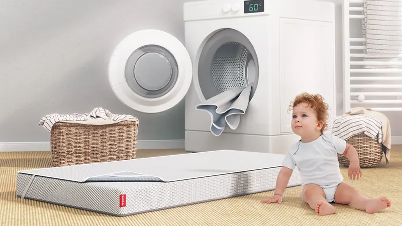 Bébé assis près d’un matelas pour enfant BODYGUARD®, devant une machine à laver ouverte dans une buanderie.