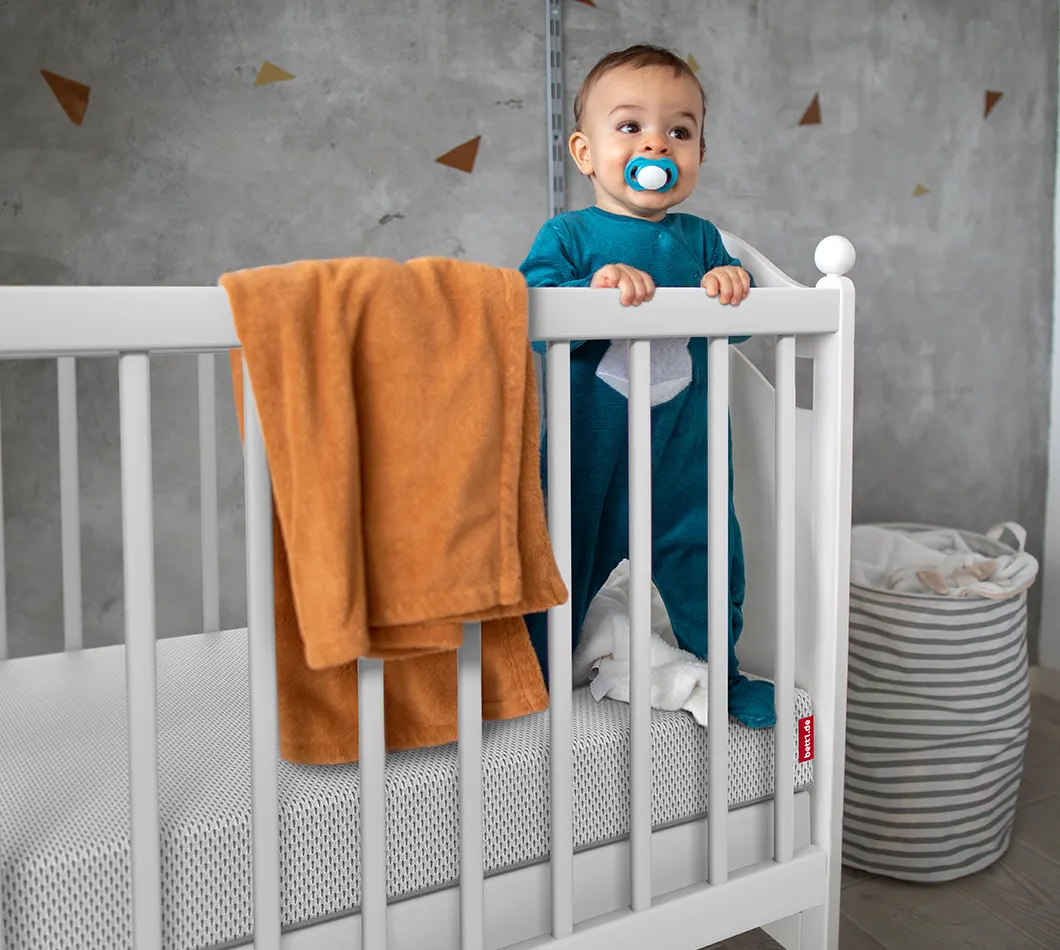 Bébé debout dans un lit à barreaux, tenant le bord. Un matelas BODYGUARD® est visible avec une housse renforcée. Un encart indique que les côtés du matelas assurent plus de sécurité lors des premiers déplacements.
