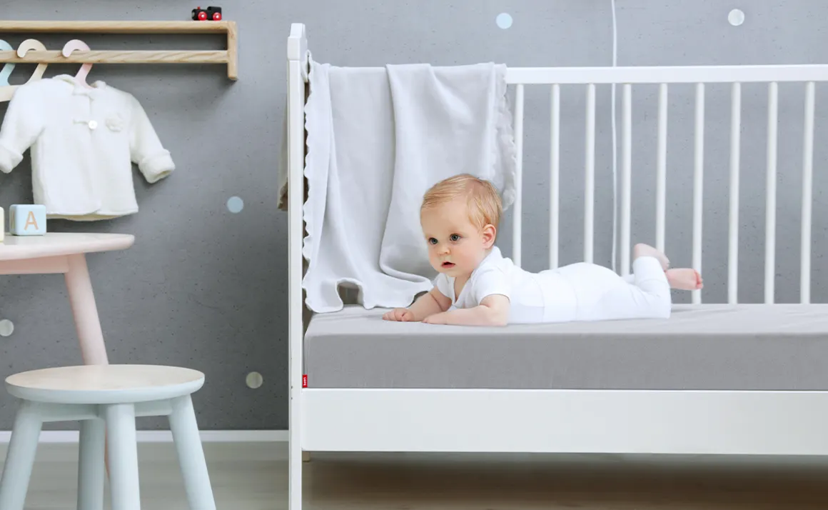 Un bébé est allongé sur le ventre la tête redressée sur un lit à barreau. Le matelas BODYGUARD enfant est recouvert du drap-housse gris enfant.
