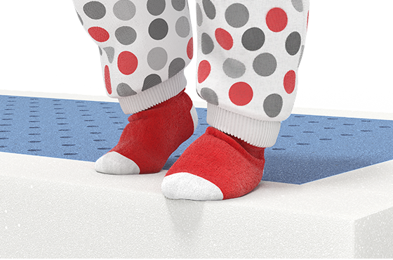Pieds d’un enfant debout sur un matelas ouvert, portant des chaussettes rouges à bout blanc et un pyjama blanc à pois gris et rouges. Les pieds sont posés sur une surface en mousse bleue perforée.