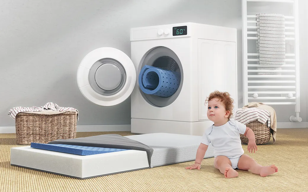 Bébé assis par terre près d'une machine à laver ouverte dans laquelle ont voit le noyau du matelas. Près du bébé, le matelas enfant BODYGUARD est déhoussé et le noyau 