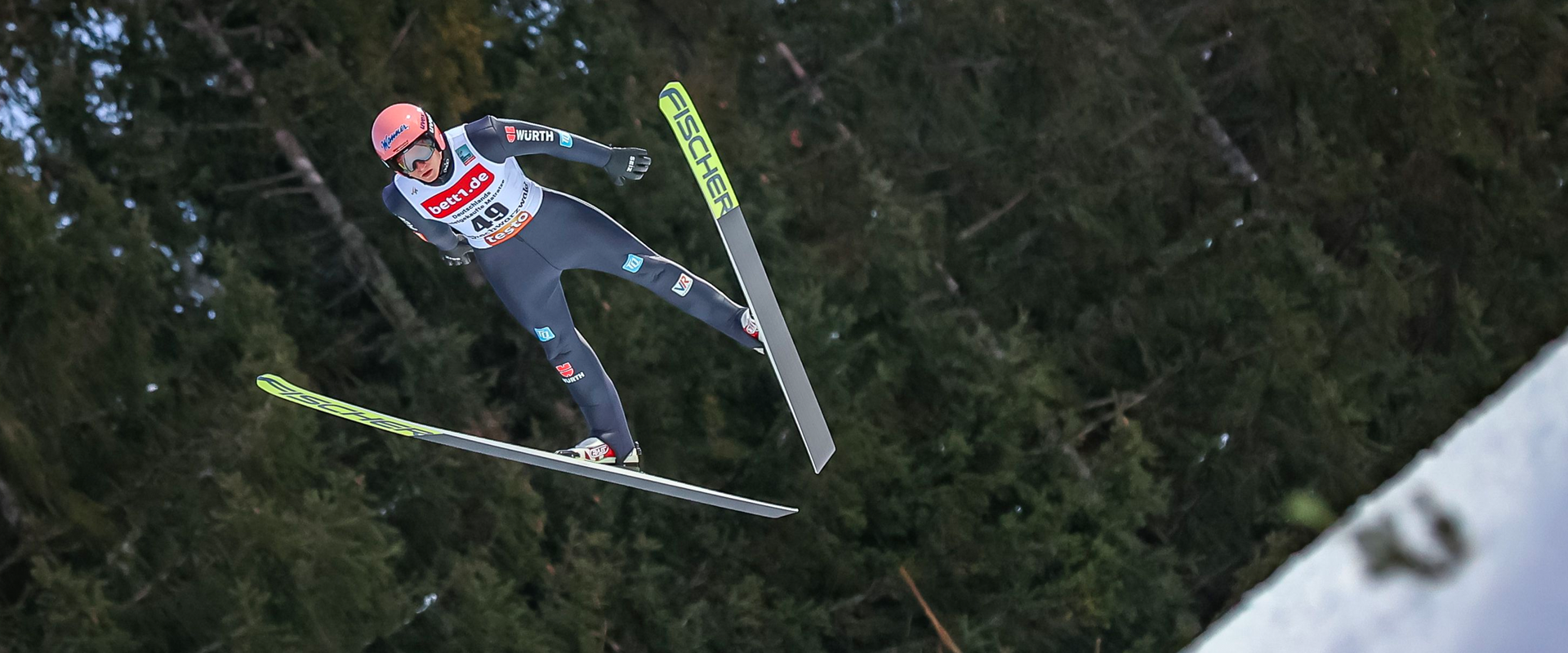 Skieur en combinaison bleue effectuant un saut à ski, vu de dos en plein vol au-dessus d’une forêt