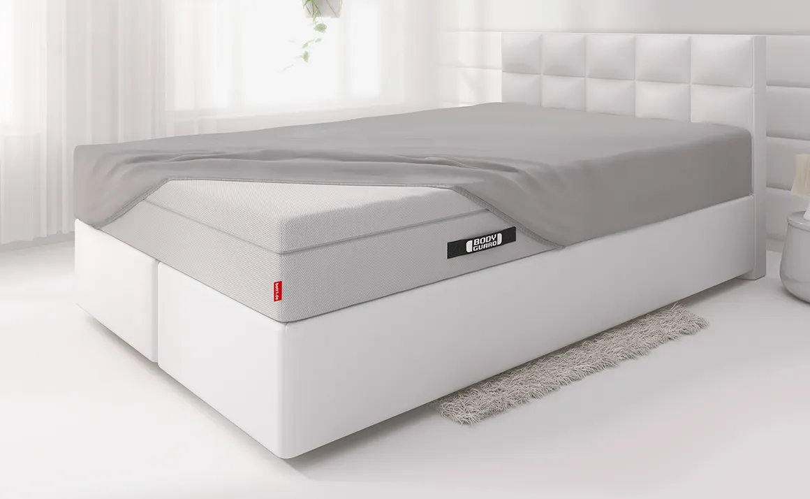 Die BODYGUARD® Boxspring Matratze ist bis auf eine Ecke mit dem Spannbettlaken extra bezogen und liegt auf einem Boxspringbett in einem hellen Schlafzimmer