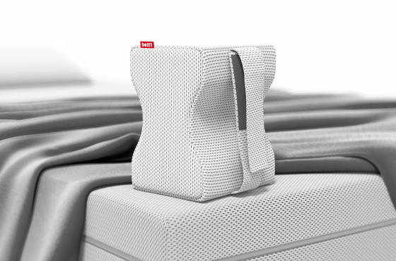 Coussin de genoux BODYGUARD® blanc à structure ergonomique, posé à la verticale sur un matelas. Il est recouvert d’une housse blanche texturée avec étiquette rouge bett1. Draps gris disposés en arrière-plan sur le lit.