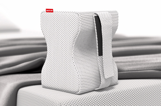 Coussin de genoux BODYGUARD® blanc à structure ergonomique, posé à la verticale sur un matelas. Il est recouvert d’une housse blanche texturée avec étiquette rouge bett1. Draps gris disposés en arrière-plan sur le lit.