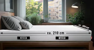Eine BODYGUARD® Matratze liegt in einem Schlafzimmer. An der Längsseite ist ein Pfeil mit "ca. 210 cm", an der Breitseite ein Pfeil mit "ca. 180 cm" beschriftet.